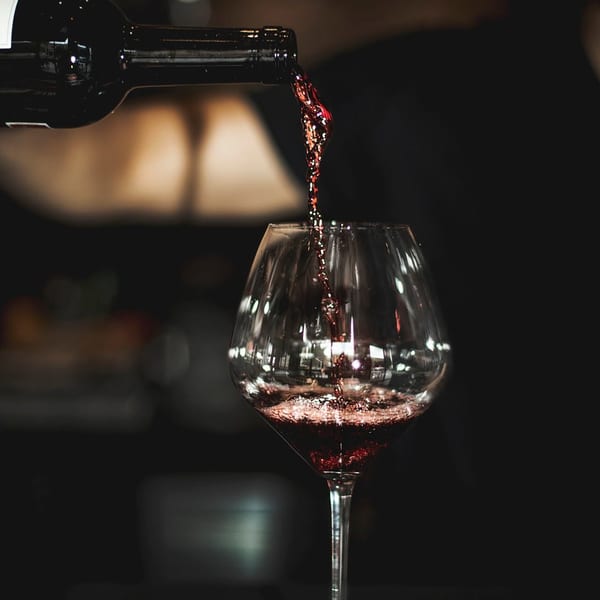 Red wine pours from a bottle into a stemmed glass against a dark background.