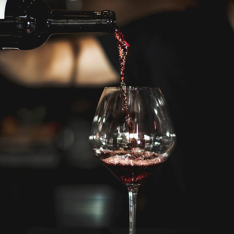 Red wine pours from a bottle into a stemmed glass against a dark background.