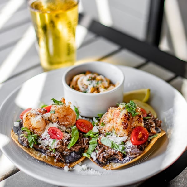 Plate of two shrimp and beef tostadas with tomatoes, cheese, and a side dish, plus a beer.