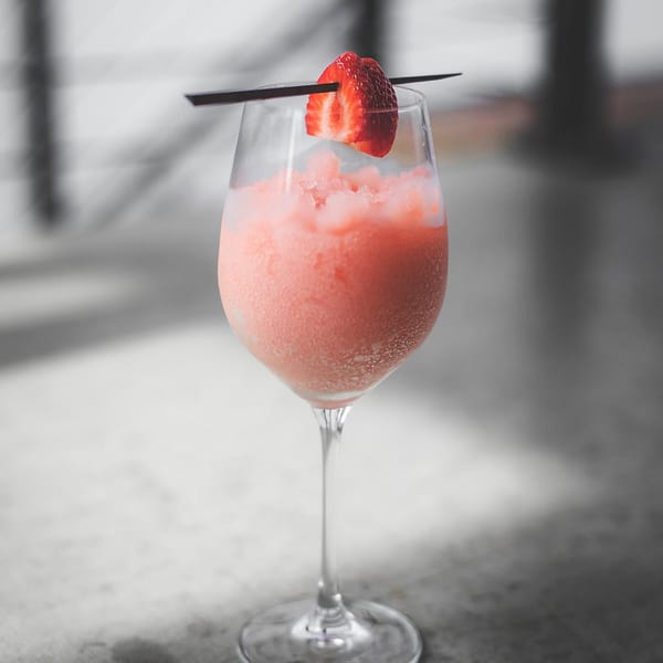 Pink frozen drink in a wine glass, garnished with a fresh strawberry half.