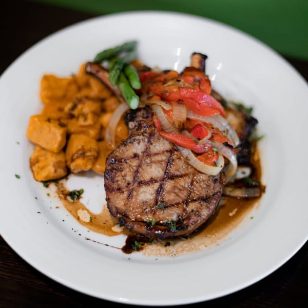 Grilled pork chop topped with peppers and onions, served with gnocchi and asparagus on a white plate.