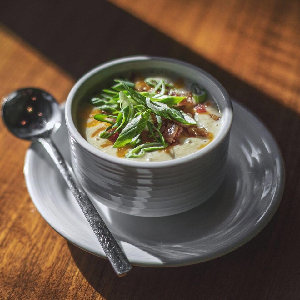 A bowl of creamy soup with green onions and bacon, next to a spoon on a wooden table.