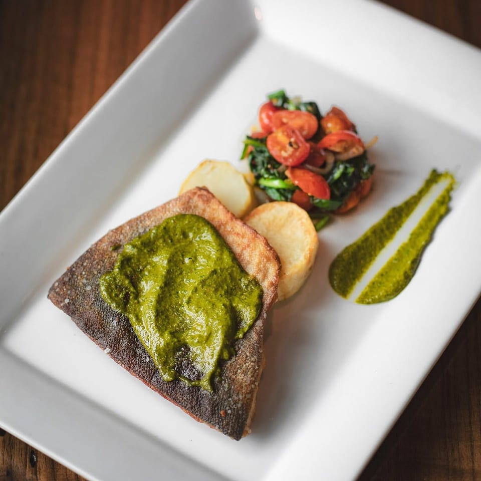 Grilled fish topped with vibrant green sauce, sliced potatoes, and a side of tomatoes and greens.