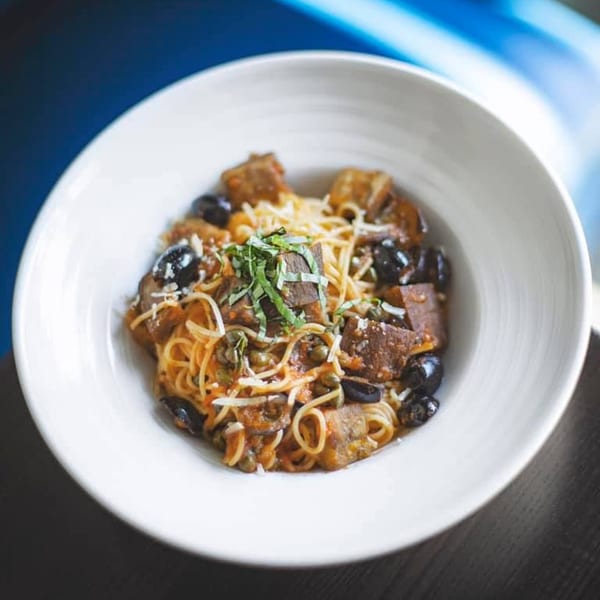 White bowl of spaghetti with braised beef, black olives, capers, and fresh basil.